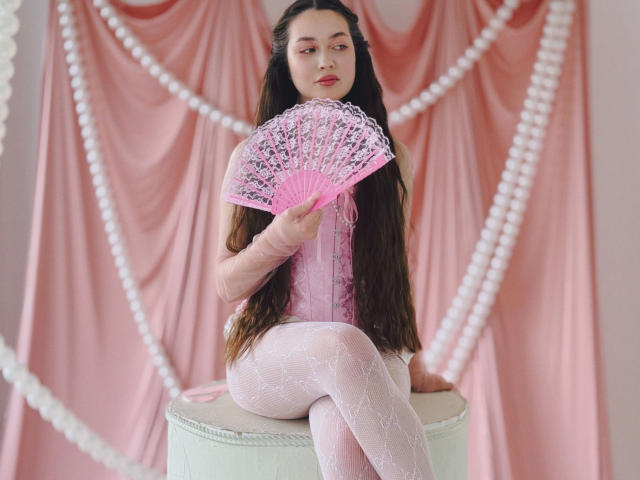 LilianCat-hot with long brown hair, pink lace corset, white patterned tights, holding pink fan, sitting on white stool, pearl strings in front, looking serious, red lipstick.
