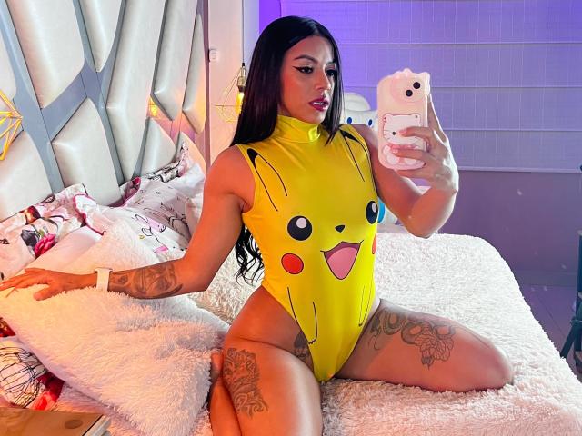 Amberv with long black hair, wearing a yellow Pikachu bodysuit, showing off tattoos on thighs and arms, kneeling on a white bed, taking a mirror selfie with a white phone.