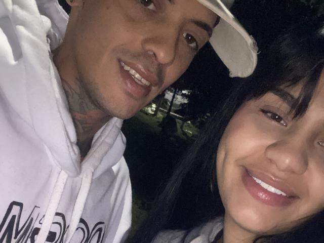 MiaWithEtan and another person, both smiling, in a selfie. MiaWithEtan has dark hair, white hoodie, and visible tattoos, wearing a white cap. The other has shiny lips.