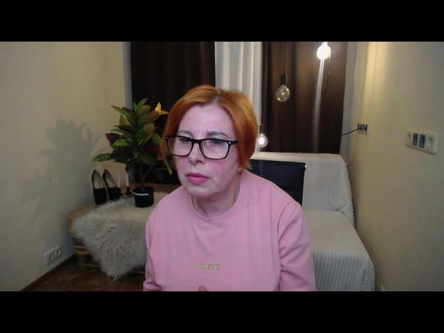 DayanaMailly, red-haired woman with glasses, pink shirt, sitting, slightly pouty face, visible white smudge on her cheek, indoors, modern room, small lamp in background.