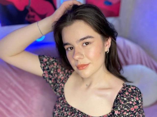 AriellaLove-hot tiene piel clara, ojos marrones y cabello oscuro. Lleva un vestido con estampado floral, está sonriendo y tiene una mano en el pelo. Está en una cama rosa, mirando a la cámara..