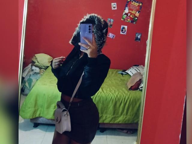 BellaOlsen with curly hair, black top, brown skirt, and gray bag, taking a mirror selfie, showing her curvy ass and small tits, in a red room with a green bed.