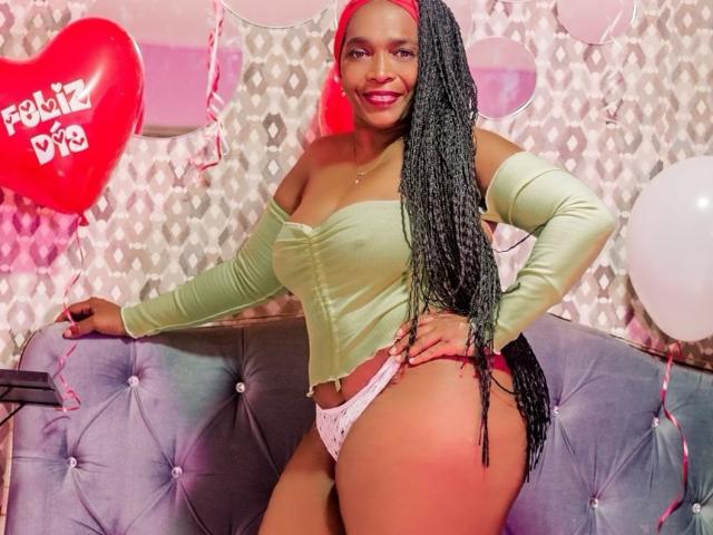 AinaEbonyQueen with long braided hair, green off-shoulder top, pink panties, big ass, smiling, pink headband, sitting on grey tufted sofa, red heart balloon, white balloons, pink string.