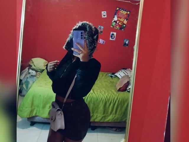 BellaOlsen with curly brown hair, black long-sleeve top, brown skirt, and beige bag, showing her curvy ass in a mirror selfie, holding a purple phone.