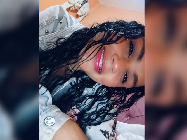 BellaOlsen has curly black hair, a cute smile with pink lipstick, and is wearing a gray shirt. Her ass is partially visible, lying on a pink pillow.