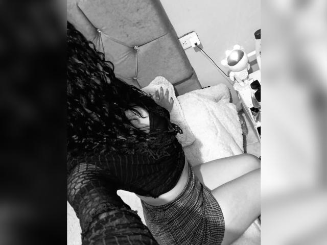 BellaOlsen with curly black hair, wearing a sheer long-sleeve top and checkered skirt, sitting on a bed, legs crossed, revealing her tits and flat stomach. Black-and-white photo.