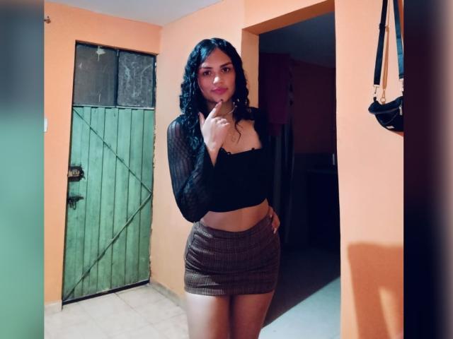 BellaOlsen with curly black hair, wearing a black sheer top and brown plaid mini-skirt, standing in a doorway, pointing at her lips with her index finger.