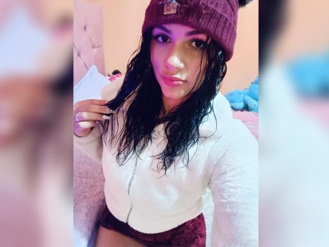 BellaOlsen with wet black hair, wearing a maroon beanie and white fuzzy hoodie, holding a paper, pink-lit face, showing her tits in maroon shorts. Soft, blurry background.