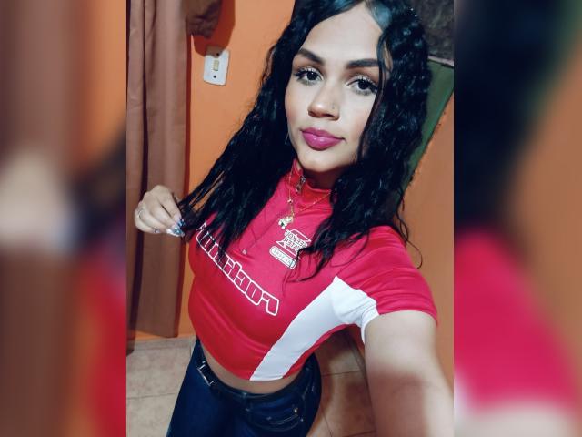 BellaOlsen with curly black hair, red crop top, and blue jeans, showing off her tits in a selfie, wearing pink lipstick, and a gold necklace in a bathroom.