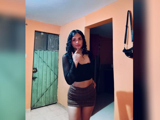 Bella Olsen mit lockigem, schwarzem Haar, die ein schwarzes Crop-Top und einen braunen Minirock trägt, zeigt mit dem Finger auf ihre Brust und steht in einem Raum mit orangefarbenen Wänden..