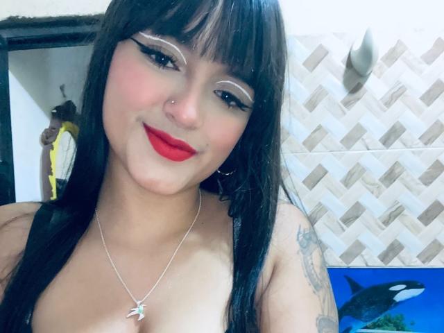 SamyWilliams hat schwarzes Haar, roten Lippenstift und Lidstrich, trägt einen Nasenring und ein Tattoo an ihrem rechten Arm, eine Kette mit einem Sternanhänger und ist obenhin nackt..
