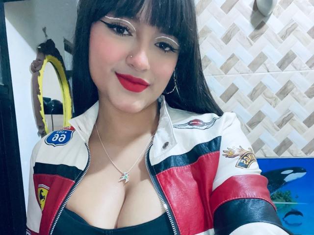 SamyWilliams mit großen Brüsten, trägt eine rote und weiße Rennjacke, ein schwarzes Oberteil, roten Lippenstift und Hula-Ohrringe, und lächelt in die Kamera. Lange, dunkle Haare, Eyeliner und ein Nasenring..