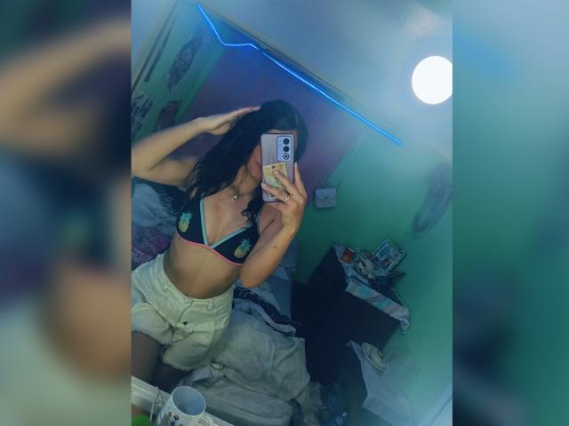 BellaOlsen with dark hair, wearing a black and red bikini top with yellow designs, and white shorts, taking a mirror selfie in a green-walled room.