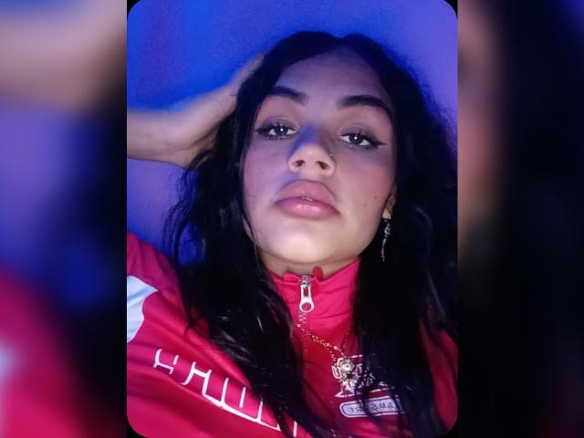 BellaOlsen with dark, wavy hair, wearing a red sports jacket, looking up with a slight pout, hand in hair, gold necklace with pendant, blue and pink lighting.