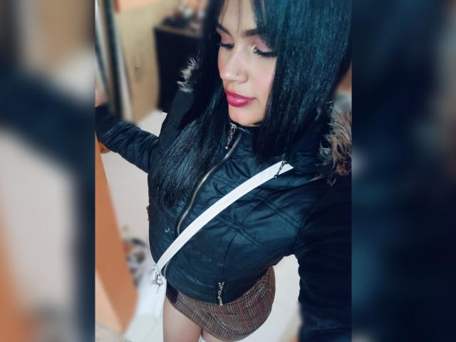 BellaOlsen with black hair, red lipstick, black puffy jacket, white strap, and brown plaid skirt, looking down, standing in a room with blurred objects.