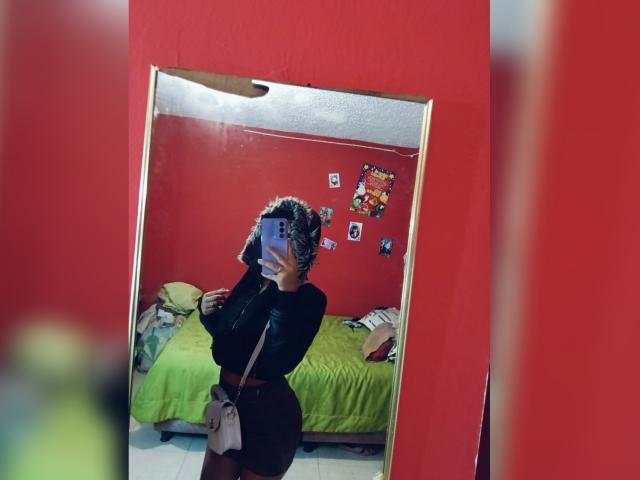 BellaOlsen, mirror selfie, red room, black long-sleeve top, brown skirt, holding phone, curly hair, small ass, green bed, pillows, wall photos, pink bag, reflection.
