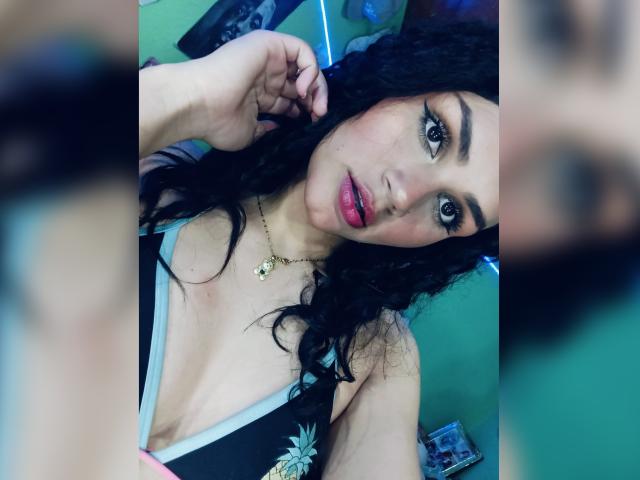 BellaOlsen with curly black hair, red lips, and green eyes, wearing a black top with a pineapple, looking at the camera, hand near head, necklace with green pendant.