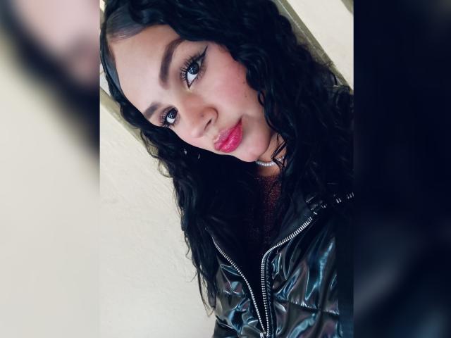 BellaOlsen with curly black hair, blue eyes, and red lipstick, wearing a shiny black zip-up jacket, looking up with a slight smile. Her tits are partially visible.