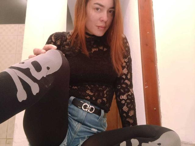PaleAndWet with red hair, black lace top, and black pantyhose with white skull patterns, sitting with one leg up, showing her black belt with a silver "G" buckle.