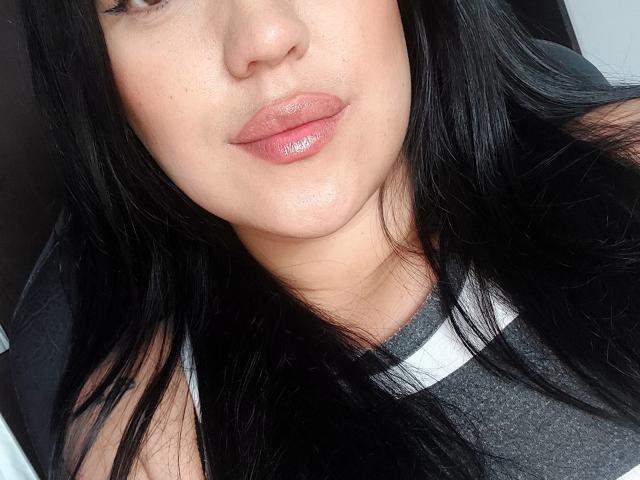 LauraRichis has shiny pink lips, light skin, and long black hair. She's wearing a gray and white shirt, with a slight sheen on her chin and upper lip.