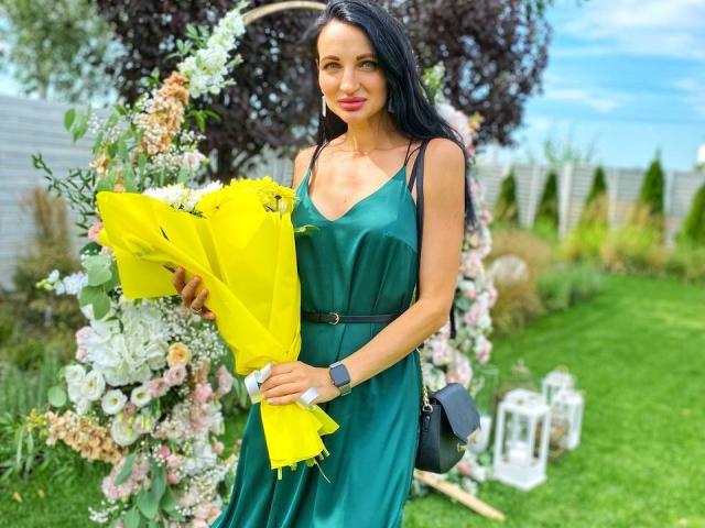 TinaMart with long black hair, green dress, holding yellow bouquet, standing in front of floral arch, wearing smartwatch, black bag, smiling, green grass, blue sky.