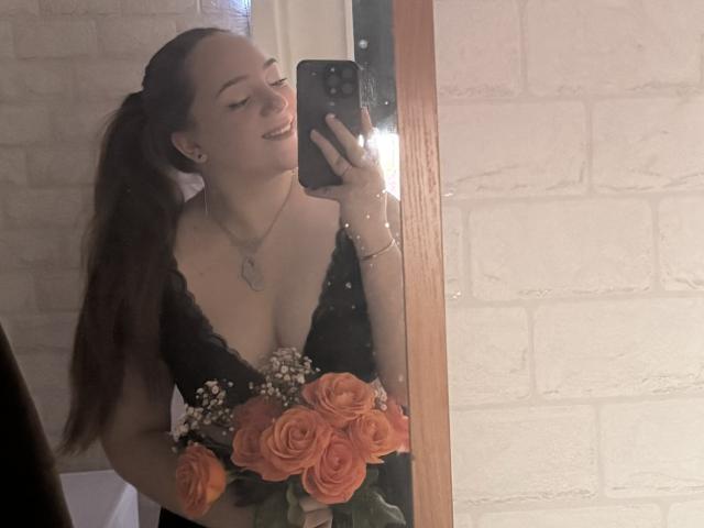 HottieFoxLalelow con una maglia nera in pizzo con rose arancioni, che tiene un telefono, sorridendo, lunga coda di capelli marroni, seno visibile, muro di mattoni bianchi, riflesso in uno specchio..
