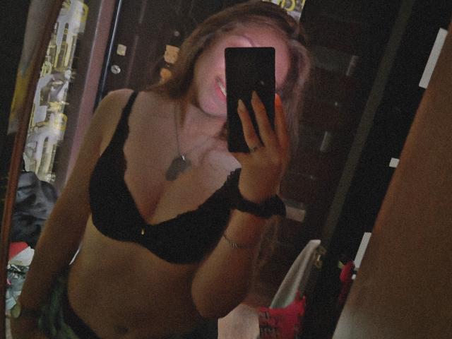 HottieFoxLalelow macht ein Selfie vor einem Spiegel, trägt einen schwarzen Spitzen-BH, zeigt kleine Brüste, lächelt und hält ein Handy in der Hand, hat braunes Haar, eine Halskette und ein Armband am Handgelenk, im schwach beleuchteten Raum..