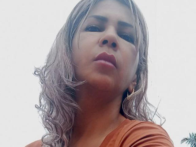 BrigiteWalsh's face in a low-angle shot, looking down with slightly parted red lips, wavy grayish-brown hair, and a brown shirt, wearing small gold hoop earrings.