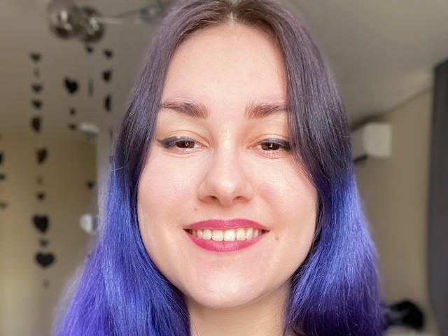 EroticSynthGoddess has purple hair, brown eyes, and is smiling with pink lipstick, showing off her teeth. Her ass is not visible, only her face is shown.