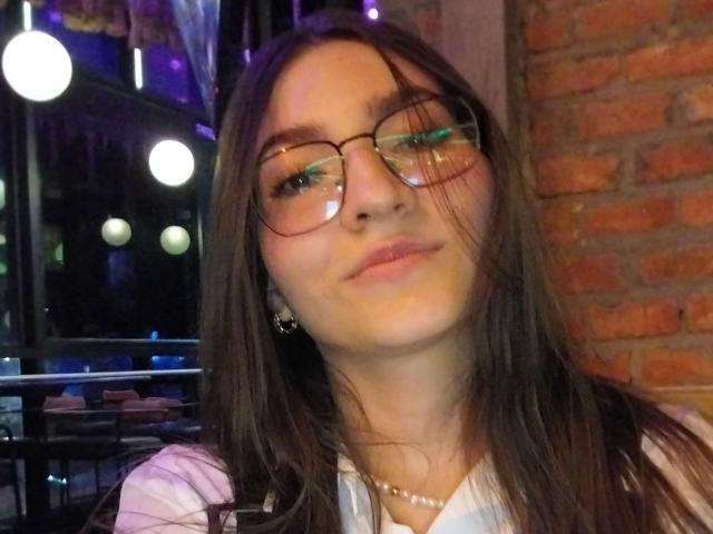 SeleneRoussel has long brown hair, green eyeshadow, and wears glasses with a subtle smile. She's in a white shirt, necklace, and has a brick wall background.