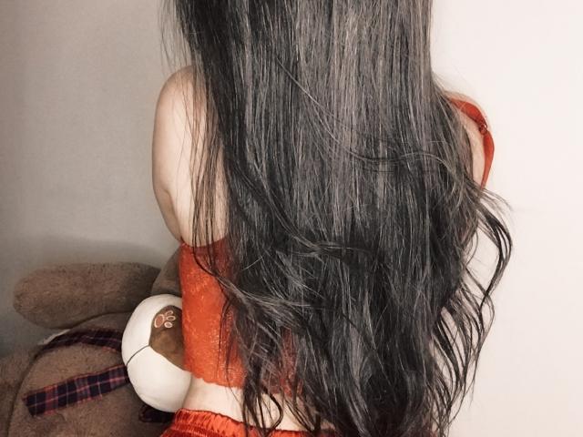 AmantaSol with long black hair, red off-shoulder top, and orange pants, sitting with a stuffed toy against a white wall. Her ass is visible.