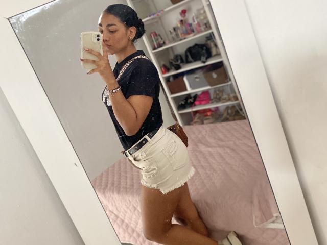 NatalyBlonder, com pele morena, camisa preta, shorts brancos de cintura alta e cinto marrom, ajoelhada, tirando uma selfie no espelho, mostrando seu traseiro, em um quarto com colcha rosa..
