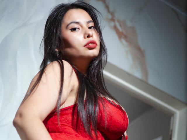 LunaMorelli with long black hair, red lipstick, and red lace dress, showing her tits, looking down with a confident, sexy expression, in a well-lit, slightly angled shot.