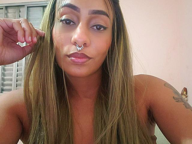 JulietCarter has long brown hair, a nose ring, and winged eyeliner. She's topless with a tattoo on her right arm, looking at the camera with a slight smile.