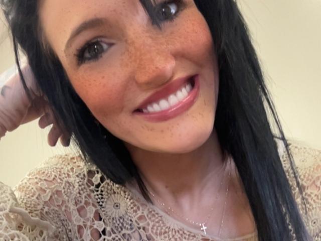XPeachesNcreamX has freckled, smiling face, black hair, white lace top, and a silver cross necklace. She's posing with hand near head, showing white teeth and pink lips.