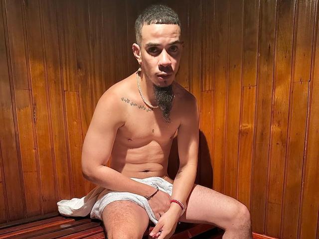 WildLeoHot is a shirtless, hairy dude with a beard, sitting on a wooden bench, covering his head with one hand, wearing white shorts, and looking down.
