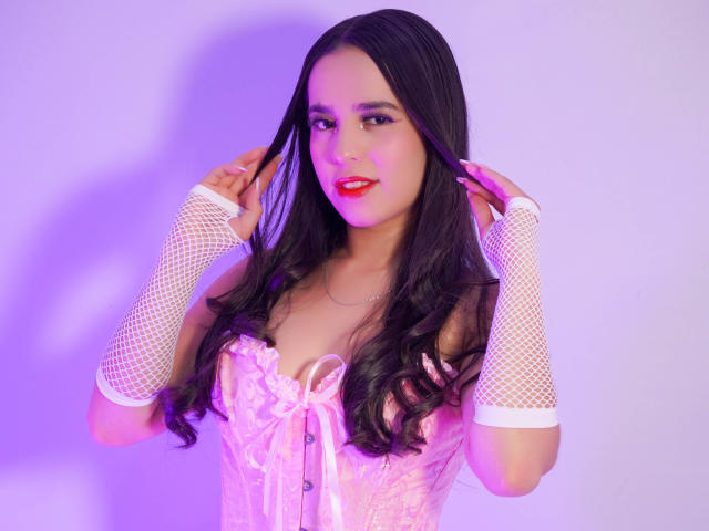 EmmaLatin, dark-skinned, smiling, wearing red fishnet lingerie, arm sleeves, and stockings, lying on white bed, legs up, showing her tits and ass, brown hair, wooden floor.