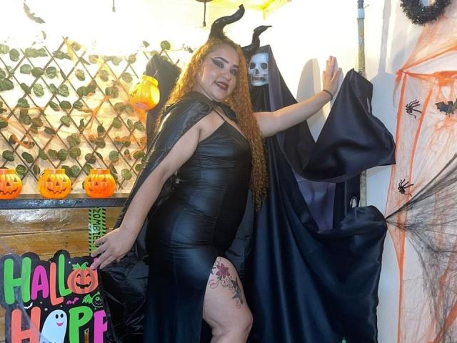SexyAndDirtyPub with long red hair in a black leather dress, showing a thigh tattoo, wearing a black cape with skull hood, holding it open, standing in a Halloween setup.