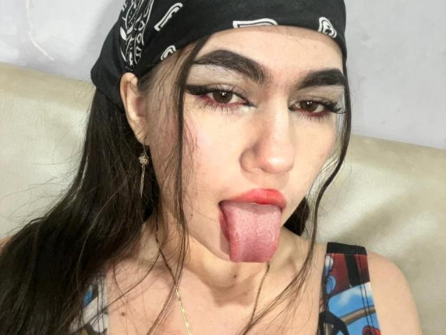 SexyAndDirtyPub with long brown hair, black bandana, and thick eyeliner, sticking out her pink tongue, wearing colorful tank top, and gold earrings, looking directly at the camera.