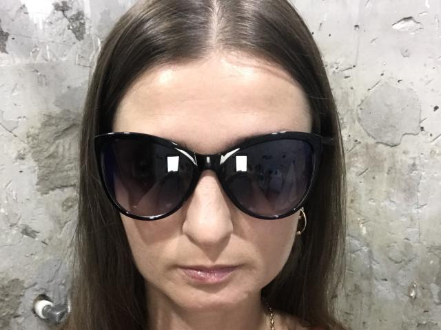 MegaBoom with straight brown hair, wearing black oversized sunglasses, has a small nose, slight smile, and gold hoop earrings, standing against a rough textured wall.