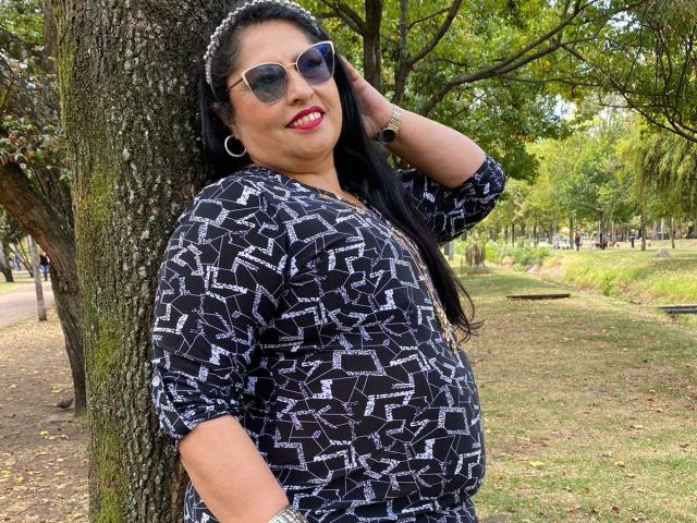 Keyla69Hot with black hair, wearing sunglasses, a black patterned shirt, and hoop earrings, standing against a tree, hand in hair, smiling, showing a slightly chubby belly.