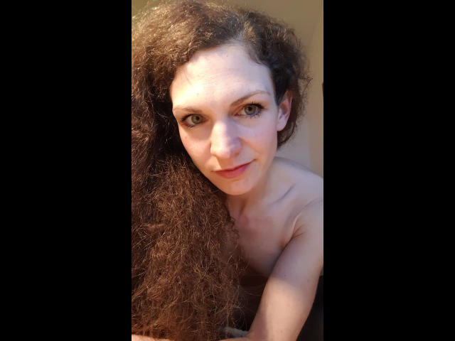 Cute-curly-hot in a red one-piece swimsuit, kneeling with one hand on her hip and the other in her curly brown hair, showing off her ass and red lips.