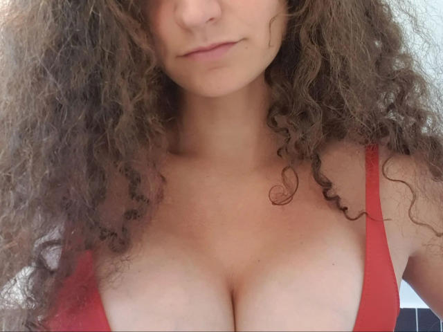 Cute-curly-hot with large, round tits and dark brown nipples, standing topless with curly brown hair, fair skin, and a neutral expression.