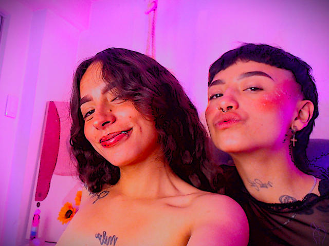 RileyAndAlexa con i capelli neri corti, un anello al naso e una giacca bianca, accanto a RileyAndAlexa con i capelli ricci e un maglione bianco, entrambe con le labbra strette. Illuminazione rosa e viola..
