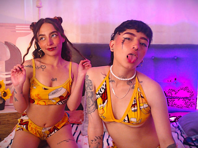 RileyAndAlexa, beiden met gele cartoon bikini's, zitten op een bed. RileyAndAlexa hebben tatoeages, kort donker haar en kijken naar de camera. RileyAndAlexa hebben kleine borsten..
