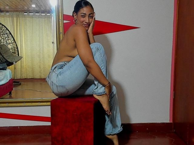 NatalyBlonder is topless, sitting on a red box, wearing blue jeans and no shoes, with her ass slightly exposed, looking back with a smile. Her tits are small.