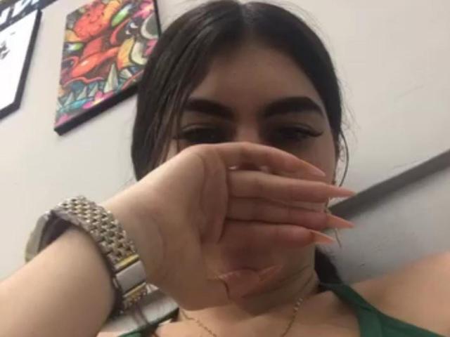 SexAndDyrtyPub with dark hair, covering her mouth with her hand, showing a cut on her lower lip. She's wearing a green top and a studded wristwatch.