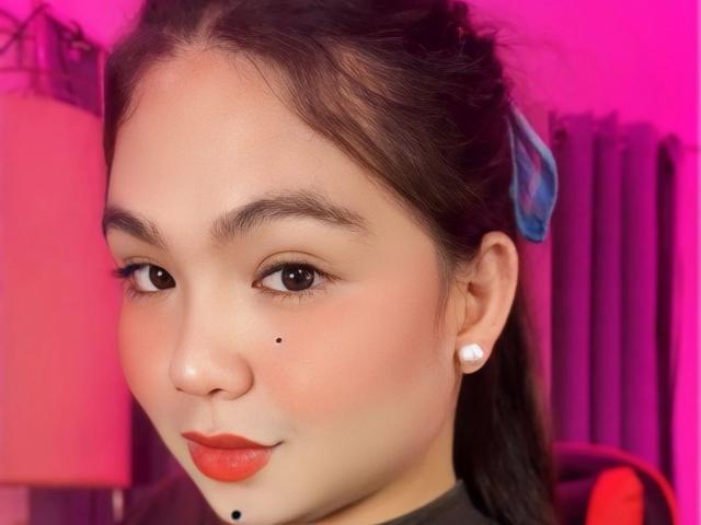 xChabelitaForYou-hot has fair skin, dark brown hair in a ponytail with a blue ribbon, brown eyes, orange lipstick, a black mole on her right cheek, and pearl earrings.
