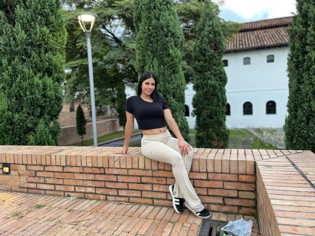 AmaiaCooper sits on a brick wall, wearing a black crop top, beige pants, and black sneakers with white stripes, her black hair down, trees and a white building in background.