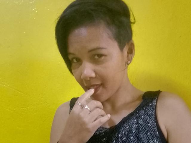 AnnaRosa69 with short black hair, wearing a sleeveless blue patterned top, touching her lips with her finger, smiling, wearing a ring, yellow background.